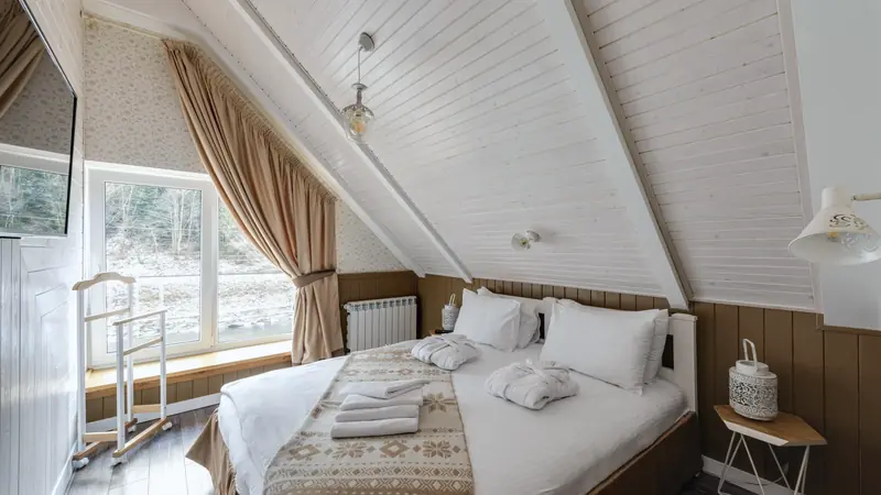 Chambre mansardée lambrissée avec un lit double aux draps blancs, sous une pente de toit et face à une grande fenêtre
