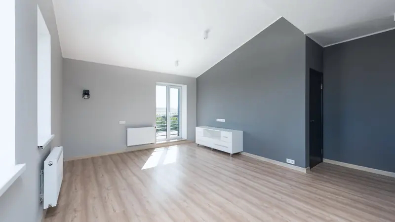 Pièce d'appartement rénové et vide, murs gris, parquet clair et porte-fenêtre ouverte sur un balcon