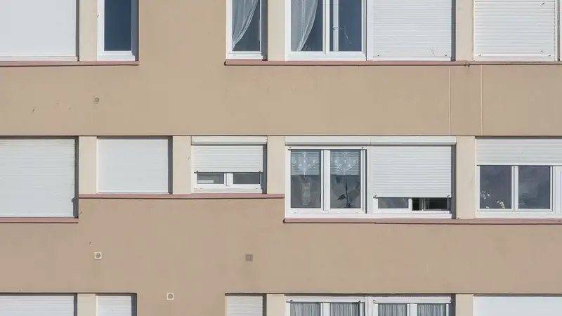 Façade beige d'un immeuble d'habitation aux fenêtres blanches et volets roulants en partie fermés