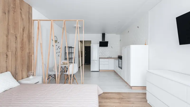 Studio meublé lumineux avec lit, claustra en bois, table pour deux et cuisine blanche en enfilade