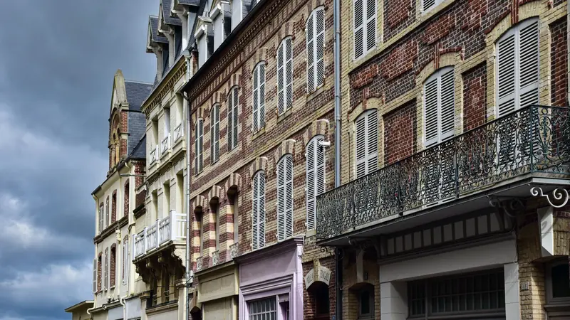 Façades d'immeubles anciens en brique et pierre à volets blancs, avec un balcon en fer forgé
