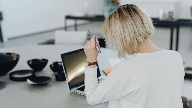 Femme vue de dos, stylo à la main, devant un ordinateur portable sur une table claire
