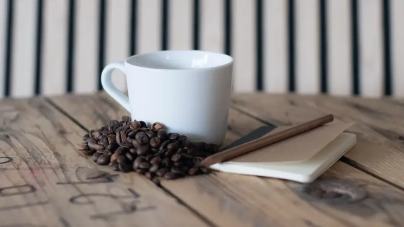 Tasse blanche, grains de café, carnet ouvert et crayon posés sur une table en bois brut