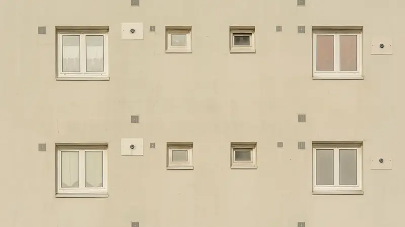 Façade crépie beige d'un petit immeuble d'habitation, fenêtres blanches alignées et grilles d'aération